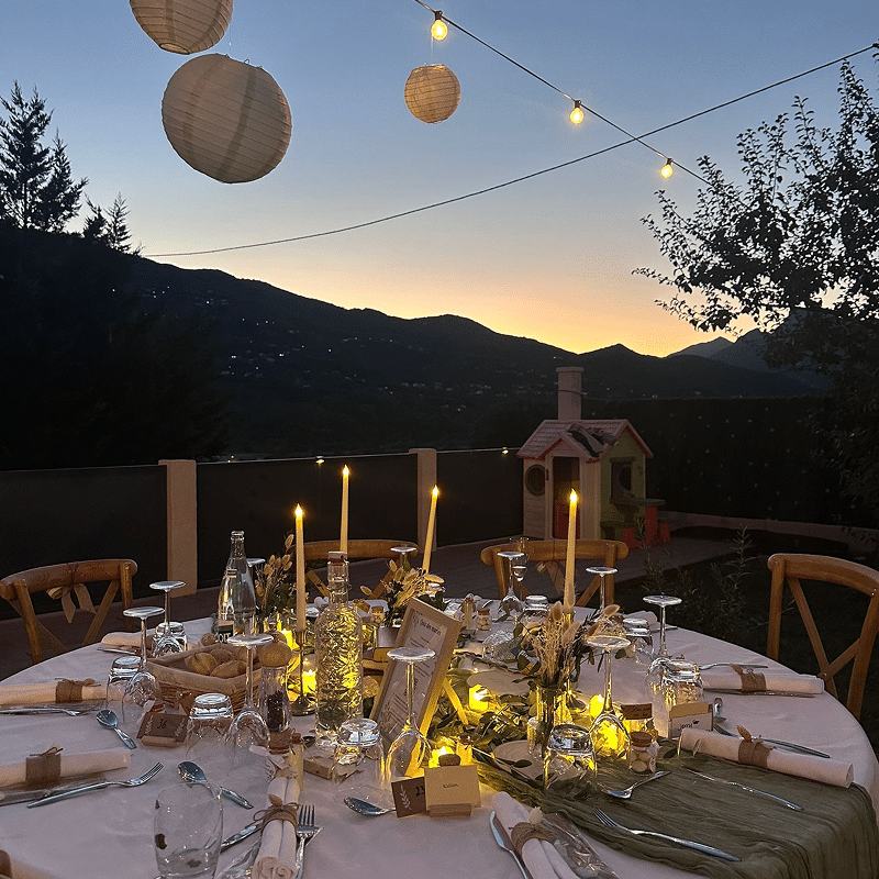 Dîner extérieur élégant au coucher du soleil, vue montagne. Table dressée avec bougies, lanternes et marque-places numérotés. Ambiance chaleureuse.