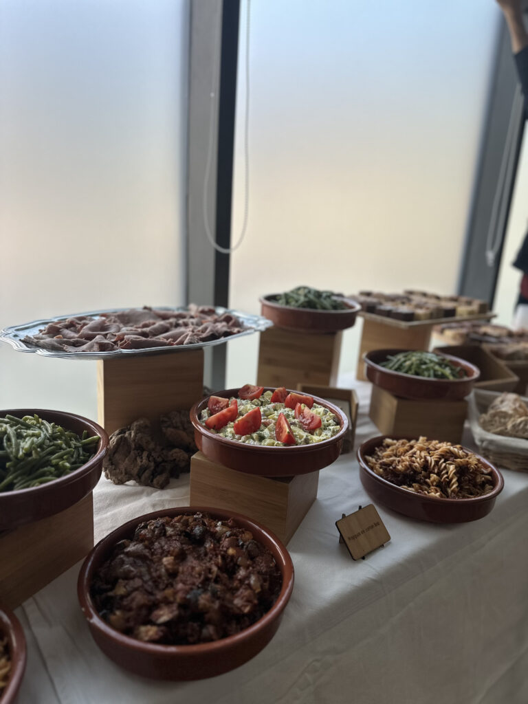 Buffet traiteur avec charcuterie en tranches fines, salade de pommes de terre aux tomates, haricots verts, pâtes à la bolognaise, et petits desserts devant une grande fenêtre lumineuse.
