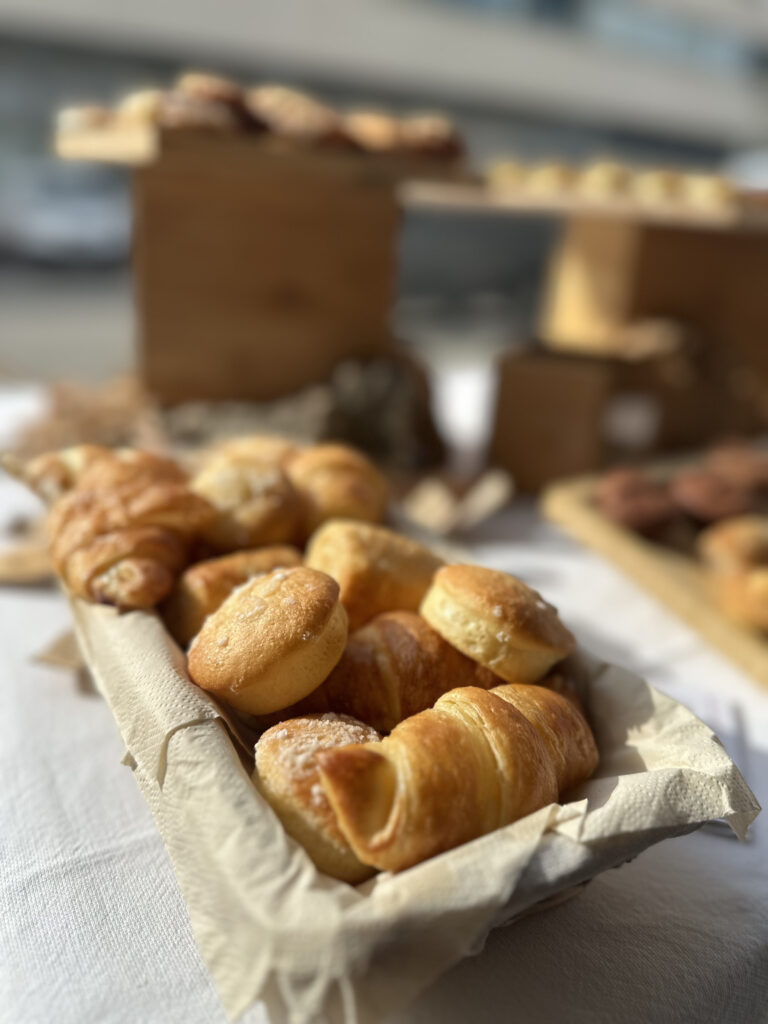 Gros plan sur des croissants dorés et des petits pains saupoudrés de sucre, présentés dans un panier tapissé de papier absorbant sur une table blanche.
