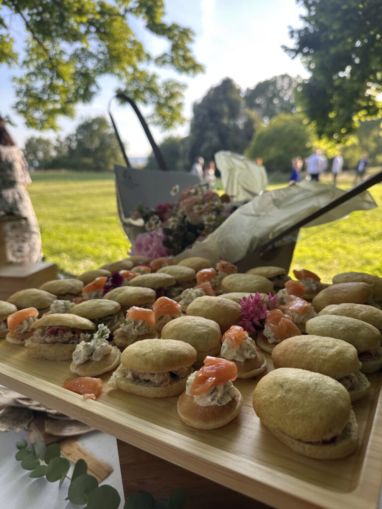Plateau de mini-sandwichs apéritifs garnis de saumon fumé et garnitures crémeuses, décorés de fleurs comestibles, en extérieur ensoleillé.