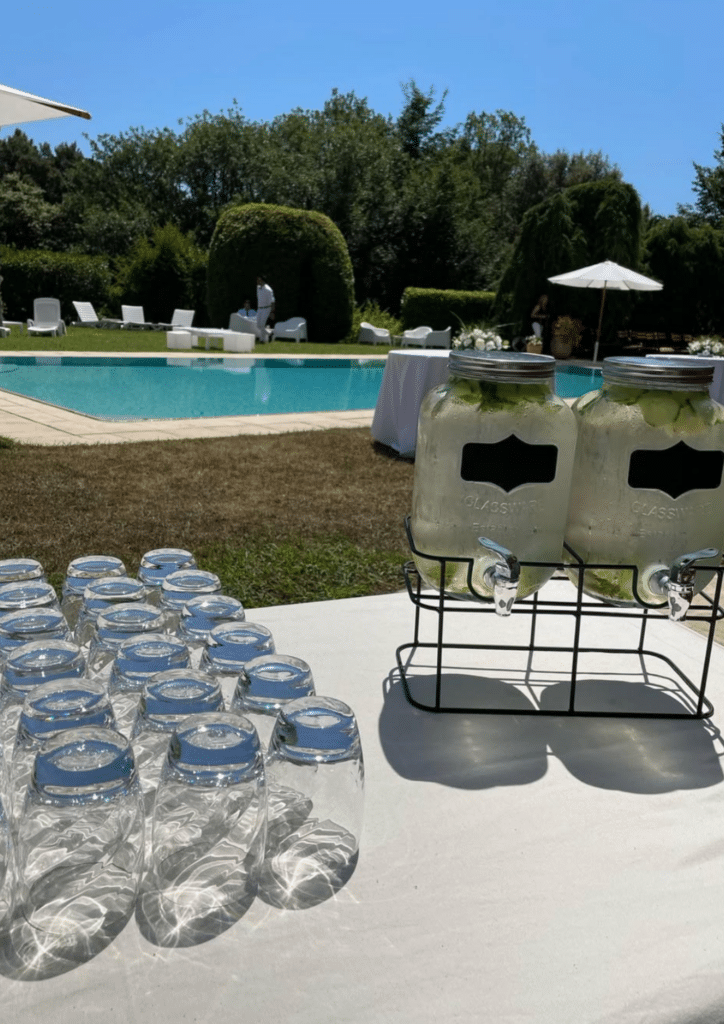 Station de Boissons pour Réception au Bord de la Piscine Verres et fontaines à eau infusée au bord d'une piscine ensoleillée lors d'une réception élégante en extérieur.