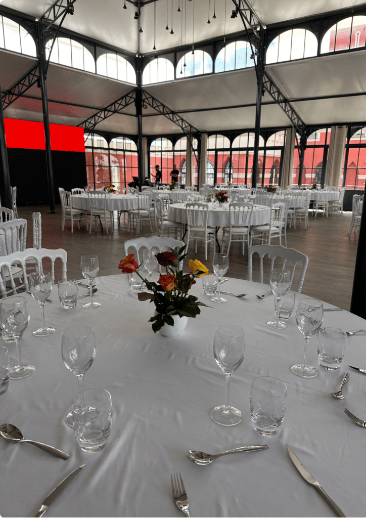 Salle de Réception de Mariage Élégante avec Tables Dressées Salle de réception élégante avec tables dressées, chaises blanches et fleurs sous une structure métallique.